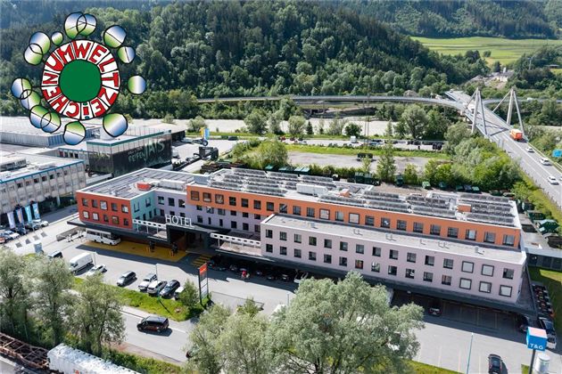A modern building with various colors, surrounded by trees and a river. In the background, hills and a bridge can be seen.