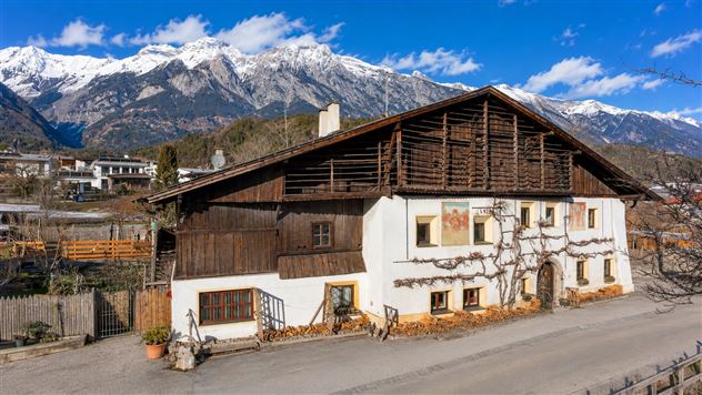 Ein traditionelles Holzhaus vor einer beeindruckenden Bergkulisse. Die schneebedeckten Gipfel kontrastieren mit dem blauen Himmel.