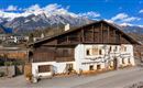 Ein traditionelles Holzhaus vor einer beeindruckenden Bergkulisse. Die schneebedeckten Gipfel kontrastieren mit dem blauen Himmel.