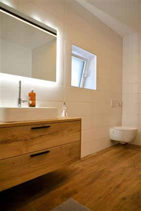 Een moderne badkamer met een houten badkamerkast, wastafel en een elegante spiegel. De kamer heeft lichte tegels en een groot raam voor natuurlijk licht.