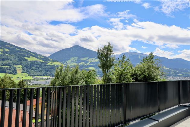 Een schilderachtig uitzicht op groene heuvels en bergen onder een heldere blauwe lucht. Op de voorgrond bevindt zich een modern balkon met een zwart leuning.