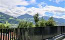 Een schilderachtig uitzicht op groene heuvels en bergen onder een heldere blauwe lucht. Op de voorgrond bevindt zich een modern balkon met een zwart leuning.