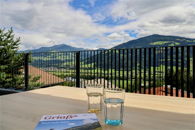 Een mooi uitzicht op groene bergen en de hemel met wolken. Op de tafel staan twee glazen water en een brochure.