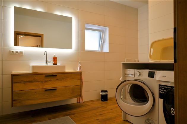 Een moderne badkamer met een wastafel, een grote spiegel en een wasmachine. De ruimte heeft heldere tegels en een warm houten ontwerp.