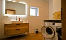 Een moderne badkamer met een wastafel, een grote spiegel en een wasmachine. De ruimte heeft heldere tegels en een warm houten ontwerp.