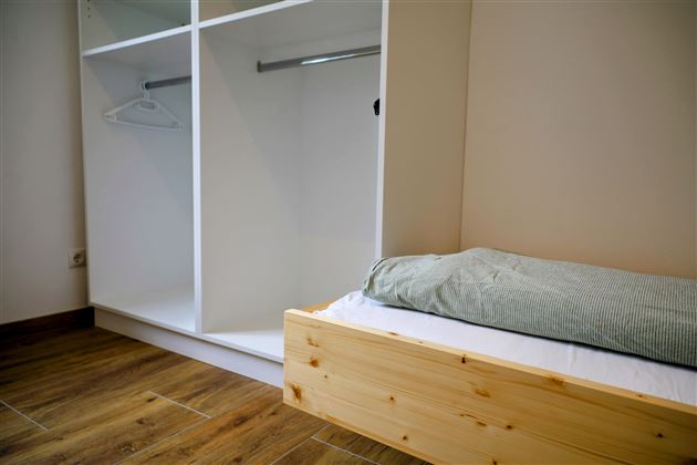 Een eenvoudige kamer met een houten bed en een open kledingkast. De muren zijn neutraal en de vloer is van hout.