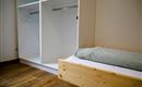 Een eenvoudige kamer met een houten bed en een open kledingkast. De muren zijn neutraal en de vloer is van hout.