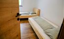 Een eenvoudige slaapkamer met twee bedden en lichte houten meubels. De muren zijn wit en er is een raam dat natuurlijk licht binnenlaat.