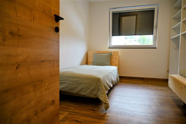 Een eenvoudige slaapkamer met een eenpersoonsbed en een groot raam. De vloer is van hout en de muren zijn in neutrale kleuren gehouden.