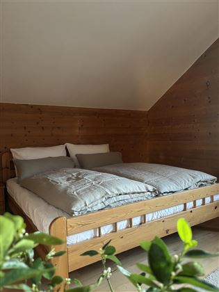 A cozy bedroom with a wooden bed and white bed linens. Plants in the foreground create a relaxed atmosphere.