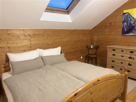 A cozy bedroom with a wooden bed and gray bedding. A colorful painting hangs on the wall, and there are a few wooden furniture pieces.