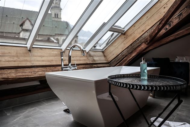 Une salle de bain moderne avec une baignoire autoportante et une belle vue à travers le velux. Au premier plan, il y a une petite table avec une bouteille.