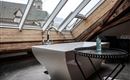 Une salle de bain moderne avec une baignoire autoportante et une belle vue à travers le velux. Au premier plan, il y a une petite table avec une bouteille.