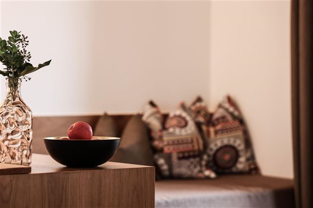 Ein stilvoll eingerichteter Raum mit einem Holztisch, auf dem eine Schüssel mit einem Apfel steht. Im Hintergrund sind gemütliche Kissen auf einer Sitzgelegenheit zu sehen.