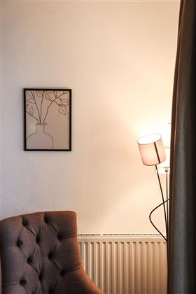 Ein gemütlicher Stuhl steht an einer Wand mit einem Bilderrahmen und einer Lampe. Der Raum hat eine warme und einladende Atmosphäre.