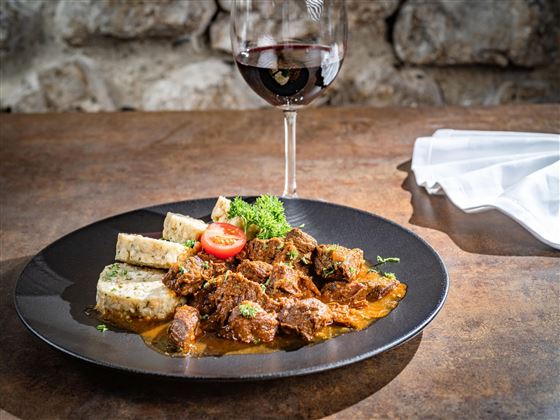 A delicious dish with tender meat in a savory sauce, served with side dishes. Accompanied by a glass of red wine on a rustic table.