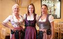 Three women in traditional Bavarian costumes stand in a cozy dining room. They smile friendly and have beer mugs on their sides.