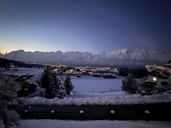 Un paysage hivernal avec des montagnes enneigées et une ville dans la vallée, qui brille dans une lumière chaude. Le ciel est clair et montre un doux crépuscule.