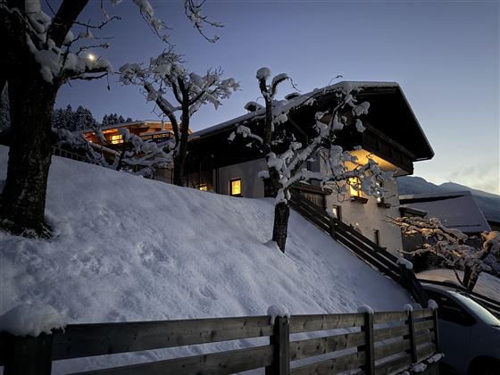Une maison confortable avec des fenêtres illuminées se trouve sous une épaisse couche de neige. L'environnement est hivernal et calme, avec des arbres recouverts de neige.