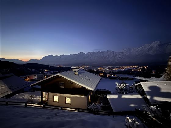 Un paysage de montagne enneigé au crépuscule. Une maison en bois avec des fenêtres éclairées se trouve au premier plan.