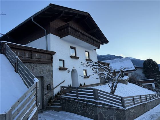 Une belle maison en style alpin, couverte de neige. L'environnement est calme et il y a des arbres et des montagnes en arrière-plan.