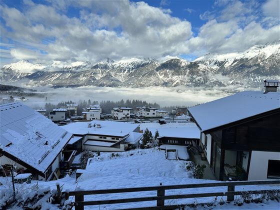 Un paysage enneigé avec des maisons et des montagnes majestueuses en arrière-plan. Le ciel est partiellement nuageux et l'atmosphère est claire et calme.