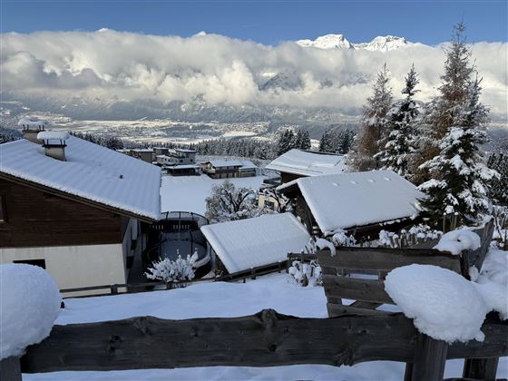 Un village de montagne enneigé avec des maisons en bois charmantes et des toits recouverts de neige. Les montagnes impressionnantes et les nuages ajoutent une atmosphère pittoresque à la scène.