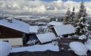 Un village de montagne enneigé avec des maisons en bois charmantes et des toits recouverts de neige. Les montagnes impressionnantes et les nuages ajoutent une atmosphère pittoresque à la scène.