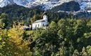 A small church stands on a hill surrounded by trees. In the background, snow-capped mountains rise beneath a blue sky.
