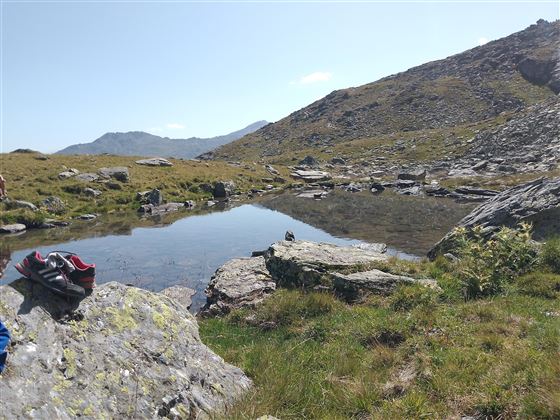 A tranquil mountain landscape with a clear pond and rocky shore. In the background, gentle hills and a blue sky are visible.