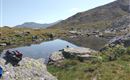 A tranquil mountain landscape with a clear pond and rocky shore. In the background, gentle hills and a blue sky are visible.