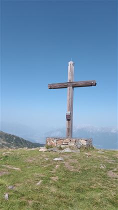 A large wooden cross stands in a meadow in the mountains. In the background, the clear blue sky is visible.