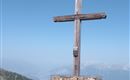 A large wooden cross stands in a meadow in the mountains. In the background, the clear blue sky is visible.