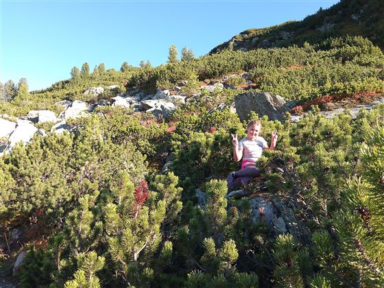 A girl is sitting in the middle of a overgrown, rocky area. She is smiling as she makes peace signs with her hands.