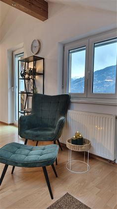 A cozy room with a green armchair and footstool. The window offers a view of the mountains and lets in a lot of daylight.