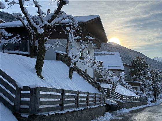 Une rue enneigée avec des maisons et un beau lever de soleil en arrière-plan. Les arbres sont couverts de neige et l'atmosphère est calme.