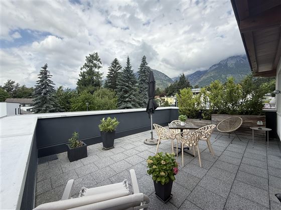 Une terrasse élégante avec des meubles modernes et une vue sur un environnement verdoyant. À l'arrière-plan, des montagnes et des collines boisées sont visibles.
