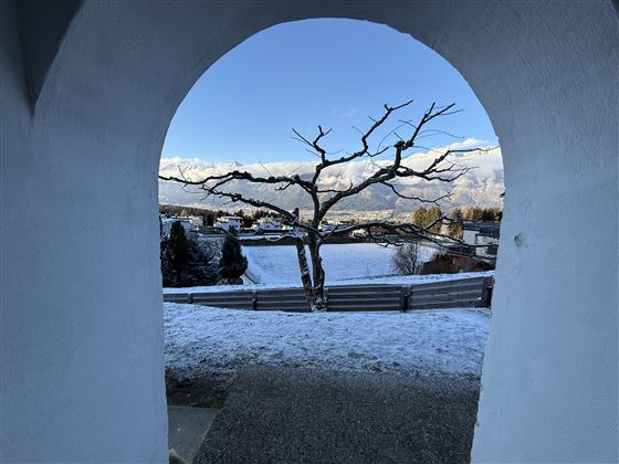 Un regard à travers un arc blanc sur un paysage enneigé. Au premier plan, un arbre dépouillé se dresse, derrière se trouvent des collines et un ciel bleu.