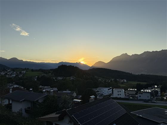 Un coucher de soleil pittoresque derrière les montagnes, qui illumine le ciel de couleurs douces. Au premier plan, des quartiers résidentiels paisibles et des champs verts sont visibles.