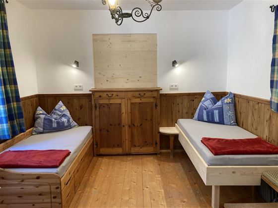 A cozy room with two beds, wooden paneling, and blue cushions. 

Checkered curtains hang on the walls, and there is a wooden wardrobe in the middle.
