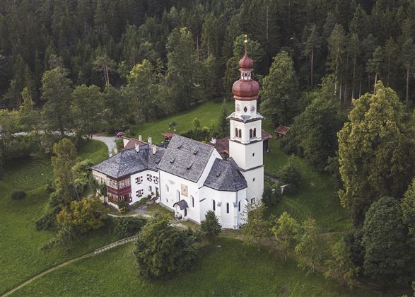 A picturesque church with a red onion dome, surrounded by green countryside and trees. The architecture features traditional elements in a peaceful setting.