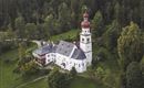 A picturesque church with a red onion dome, surrounded by green countryside and trees. The architecture features traditional elements in a peaceful setting.