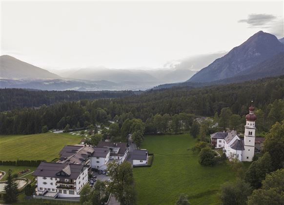 A picturesque landscape with a historical building and a church tower in the foreground. In the background, mountains and a wooded valley stretch out.
