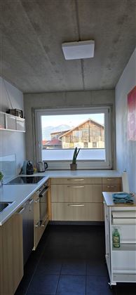 Eine moderne Küche mit Holzschränken und einem großen Fenster. Draußen sind Dächer von Gebäuden sichtbar.