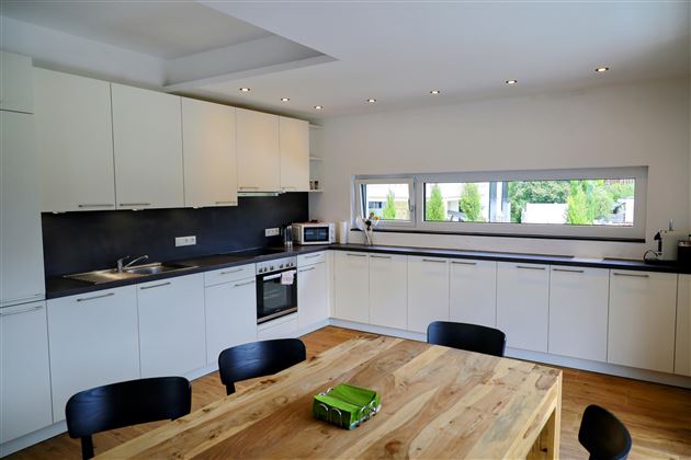 Een moderne keuken met witte kasten en een houten werkblad. Een grote tafel staat in het midden, omringd door zwarte stoelen.
