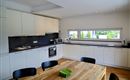 Een moderne keuken met witte kasten en een houten werkblad. Een grote tafel staat in het midden, omringd door zwarte stoelen.