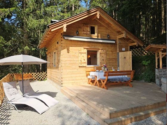 Ein gemütliches Holzhaus in der Natur mit einer Terrasse. Ein Tisch mit Stühlen und eine Liege laden zum Entspannen ein.
