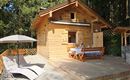 Ein gemütliches Holzhaus in der Natur mit einer Terrasse. Ein Tisch mit Stühlen und eine Liege laden zum Entspannen ein.