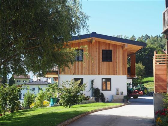 A modern house with wooden cladding and a blue roof. Surrounded by green lawn and garden plants in a rural setting.