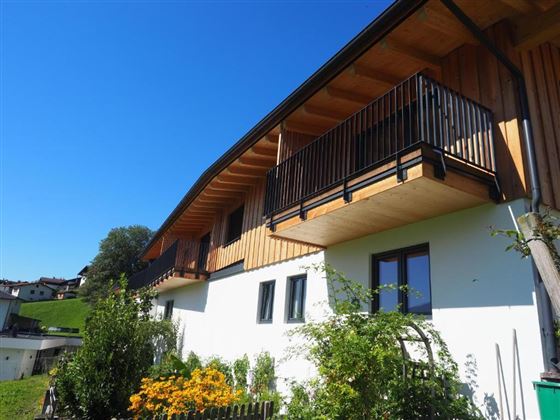 A modern, two-story house with wooden cladding and balconies. The garden is landscaped with colorful flowers and green spaces.